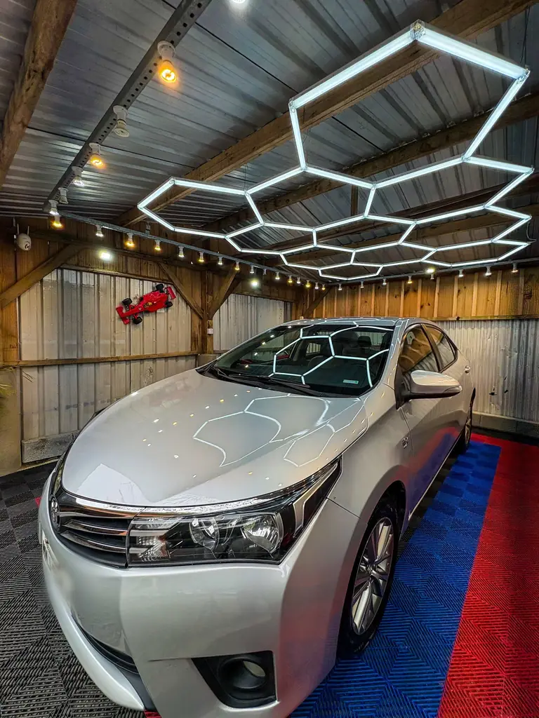 Corolla prata sob o teto hexagonal de LED, piso vermelho e azul e miniatura de Fórmula 1 na parede.