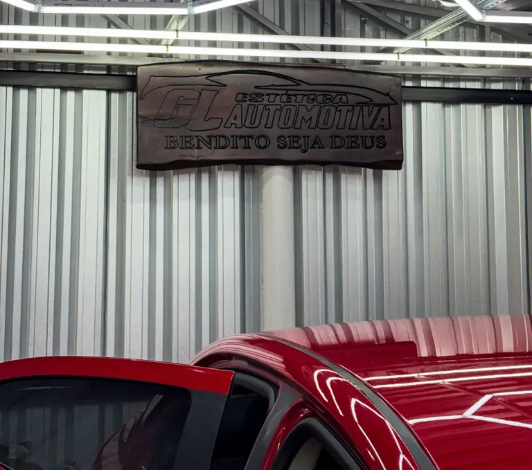Carro vermelho espelhado sob as luminárias, com a placa de madeira da GL Estética Automotiva ao fundo.